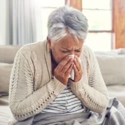 Senior woman on her couch sneezing into a tissue.